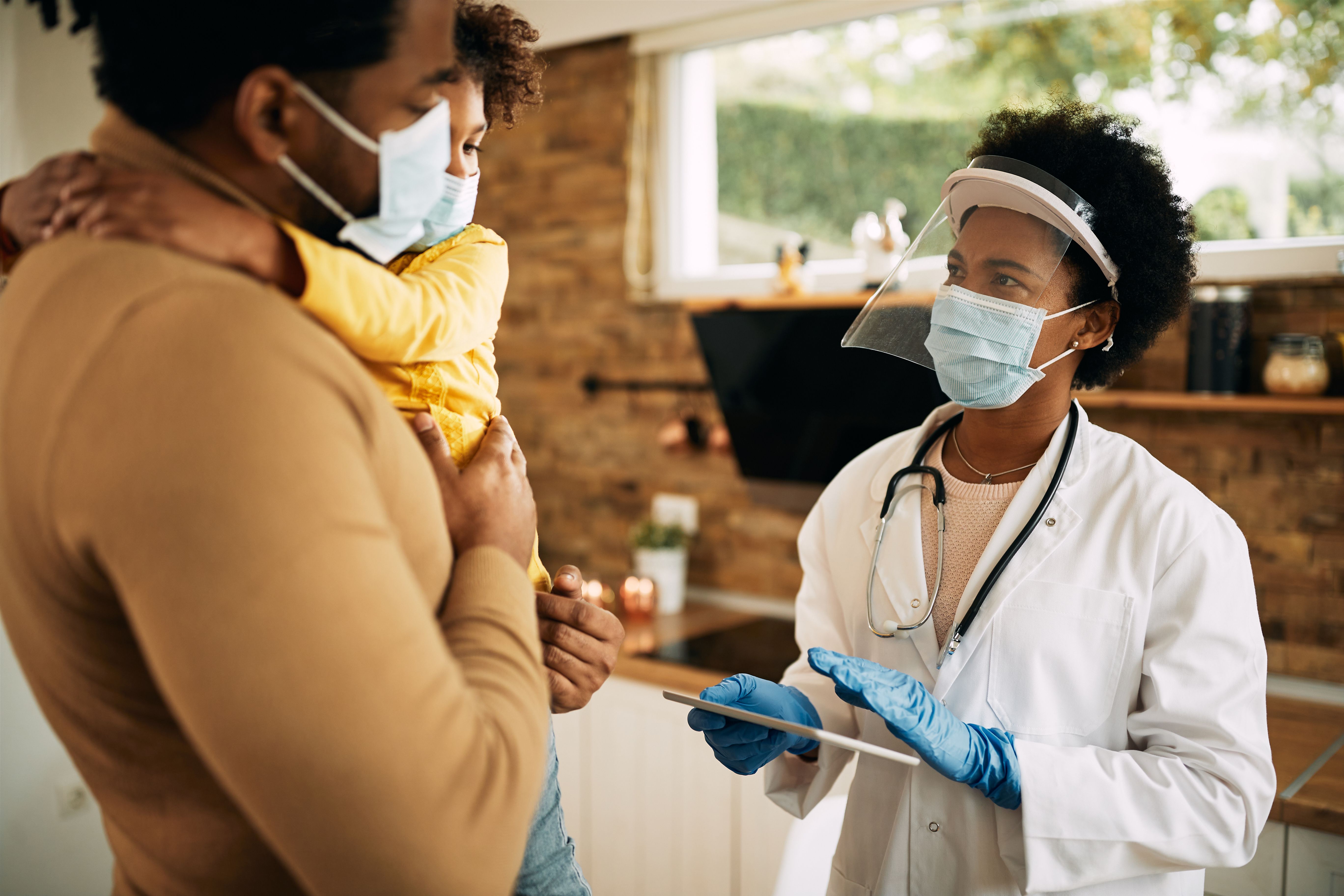 A man holding his young daughter while a doctor with a clipboard speaks to them. All are wearing masks.