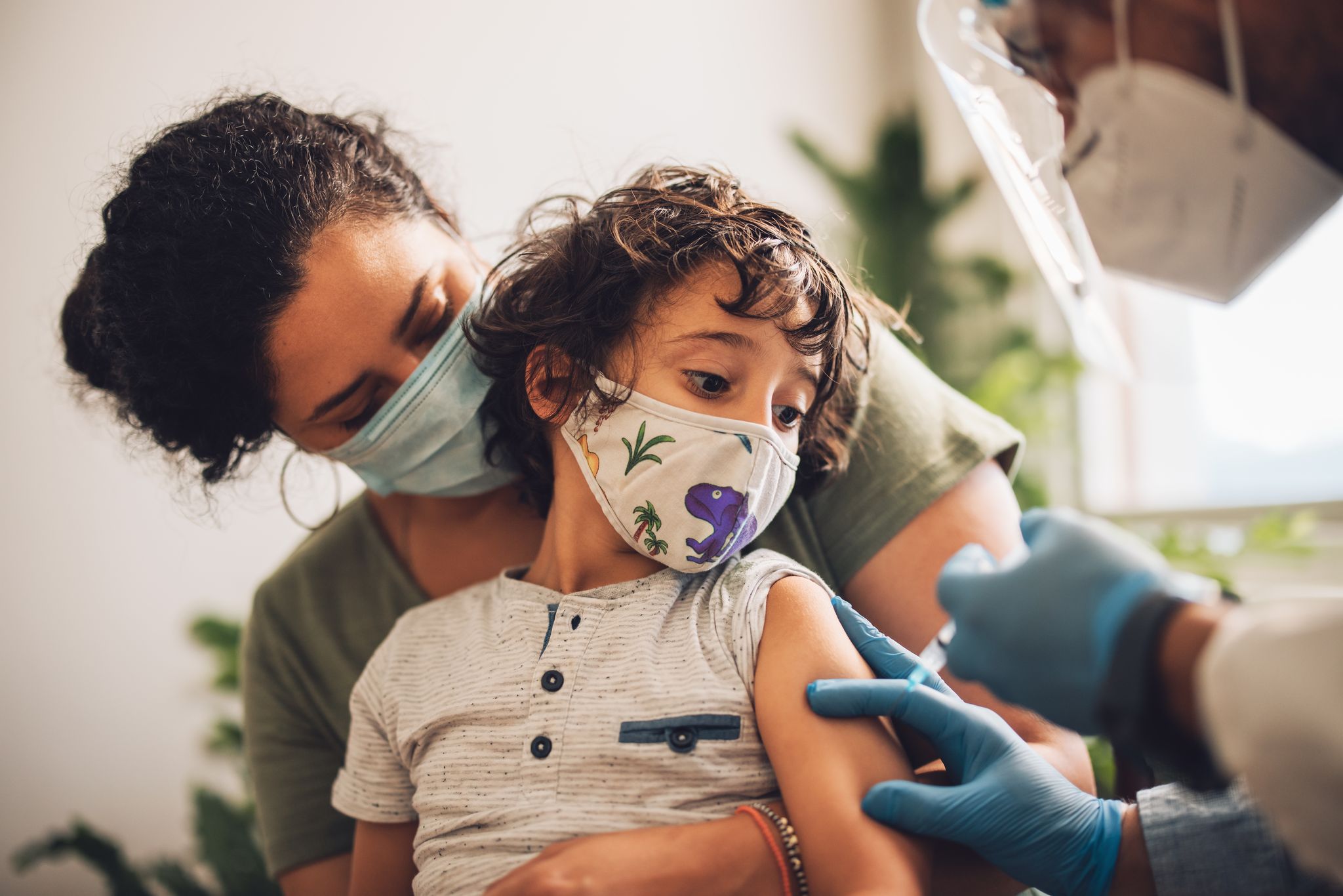 A boy wearing a mask receiving an injection from a doctor while sitting on his mother's lap.